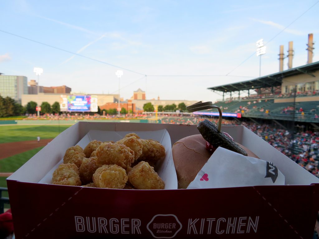 A box with tater tots on the left and a hamburger on the right in front of a baseball field.