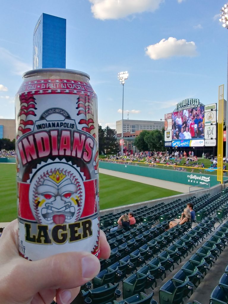 A sixteen-ounce beer can with a baseball in the artwork that reads Indians Lager in front of a baseball field with a videoboard on the right.