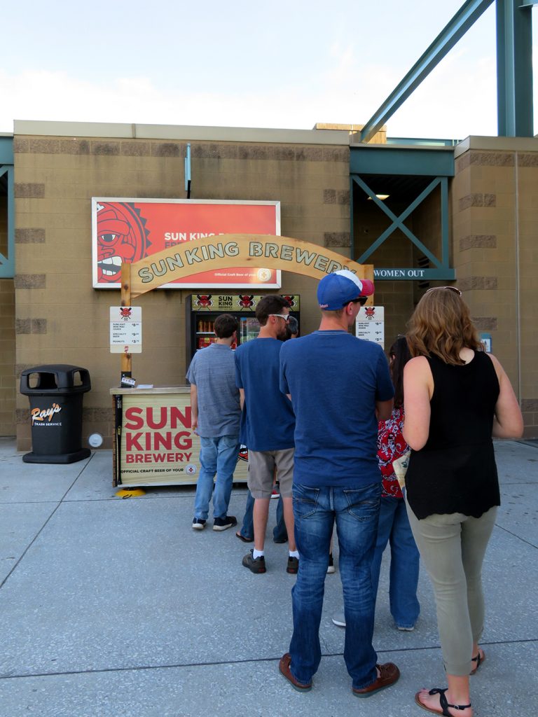 Several people stand in line at a portable concession stand selling Sun King beer.