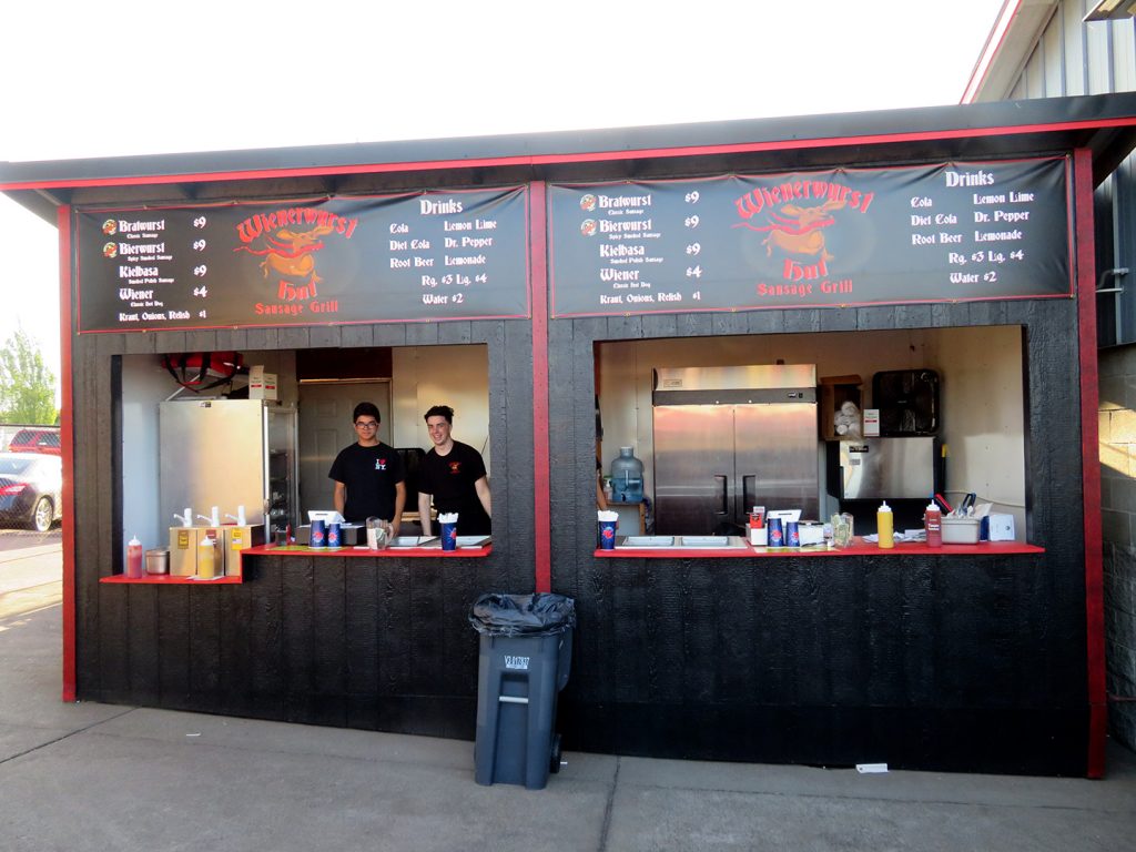 A wooden concession stand painted black with a menu above the opening and banner that says "Wienerwurst Hut."