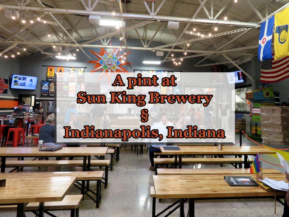 Overview of a room with several wooden picnic benches and a large bar in the background. Text overlaying image says "A pint at Sun King Brewery (Section sign) Indianapolis, Indiana."