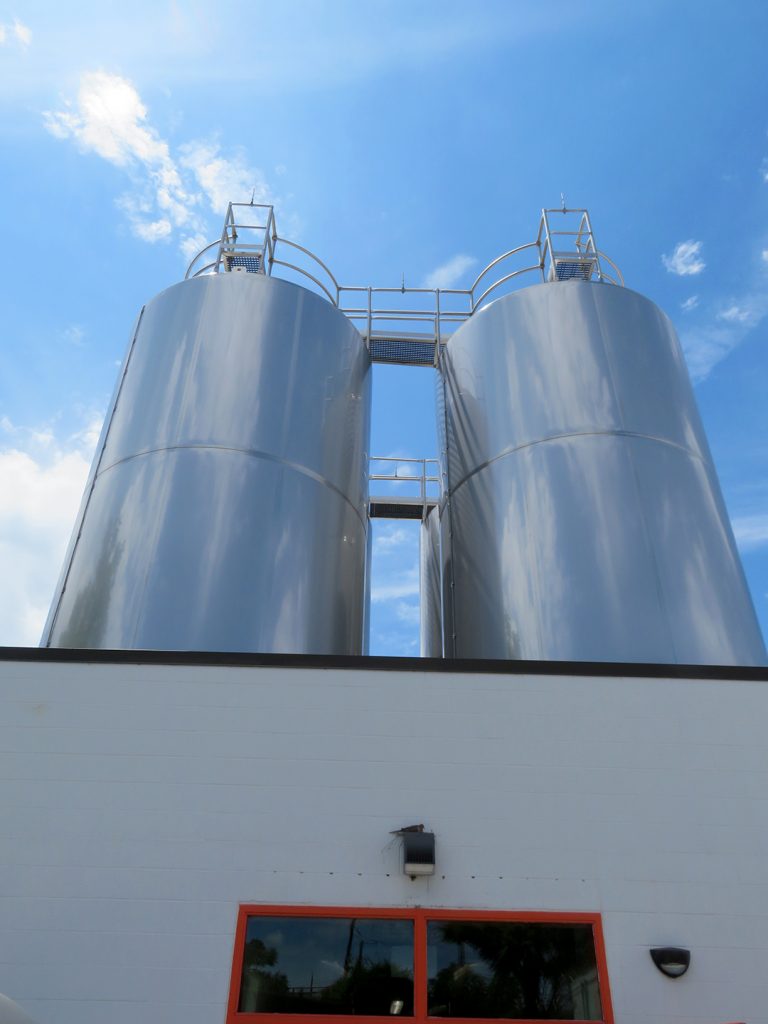 Two tall, stainless steel 420-barrel fermentation tanks tower over a cinder block building.