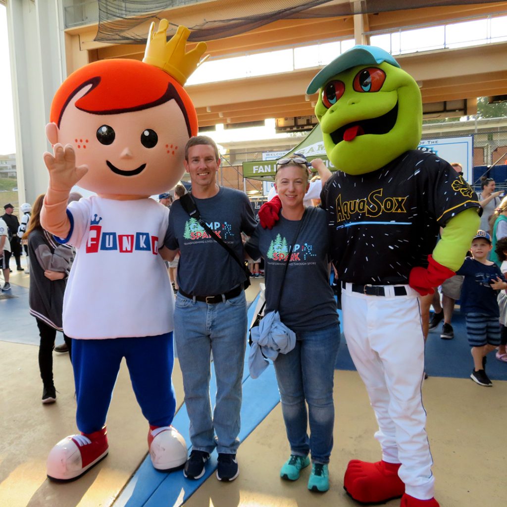 A man and woman stand between Freddy Funko, red-head human mascot for Funko, and Webbly, a frog mascot for the Everett AquaSox.
