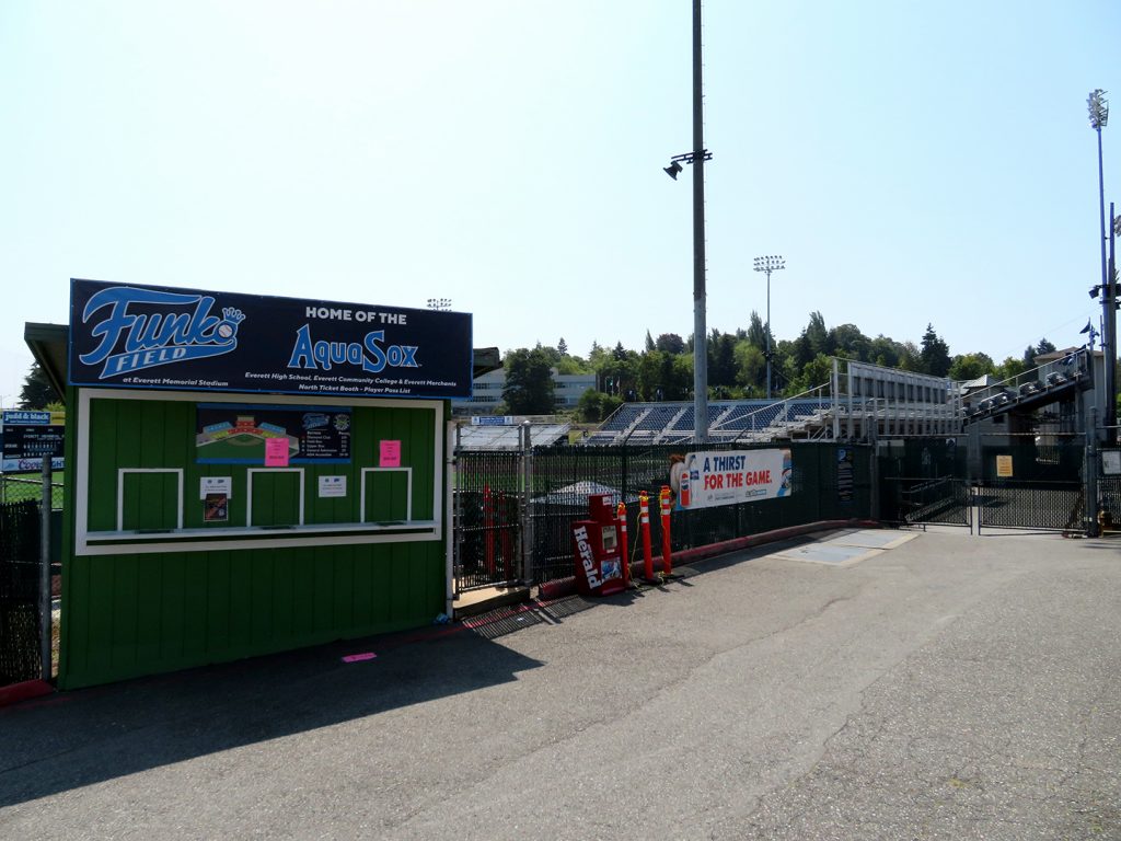 A small, wooden ticket booth with a sign above it that says "Funko Field, home of the AquaSox" and a baseball stadium behind it.