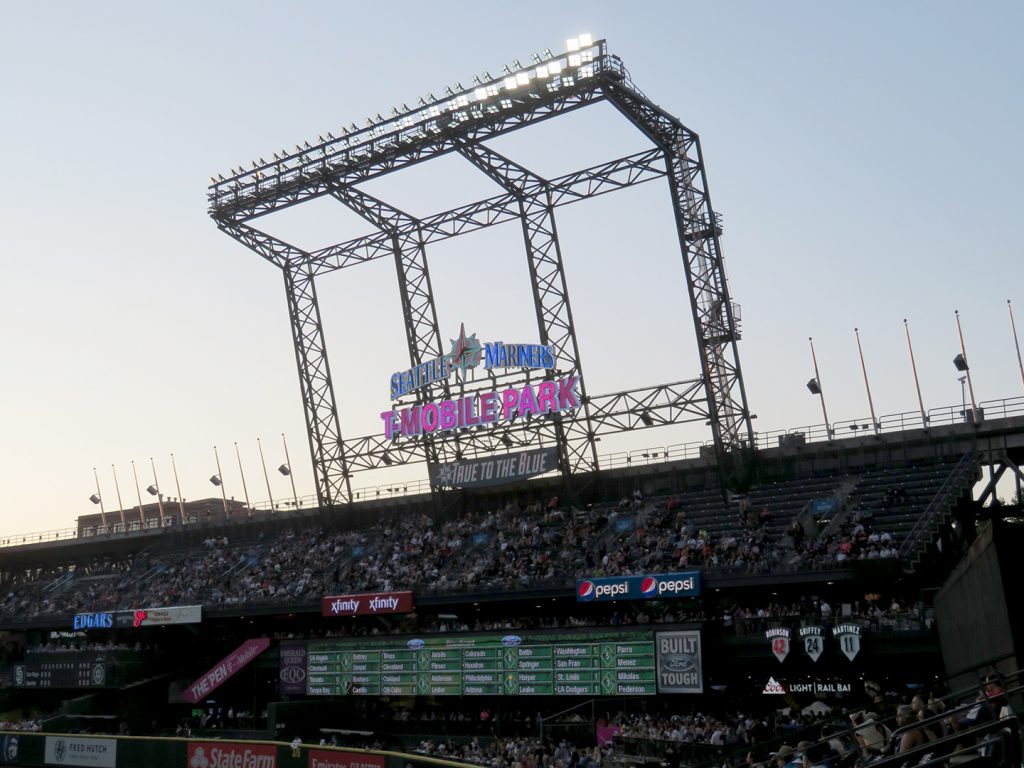 "Seattle Mariners" sign sits above "T-Mobile Park" sign with decks of seats underneath them and an out-of-town scoreboard.