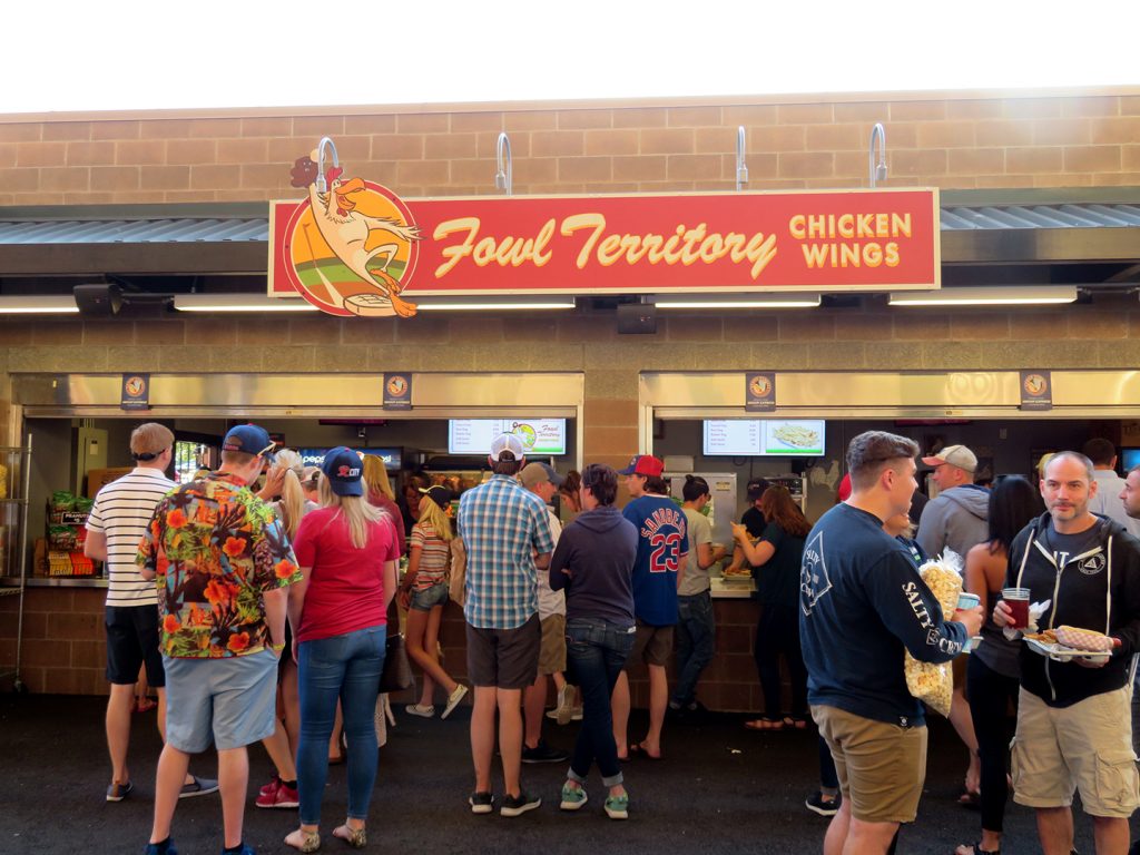 A concession stand with a sign above it saying "Fowl Territory Chicken Wings" and several people standing in front of it.