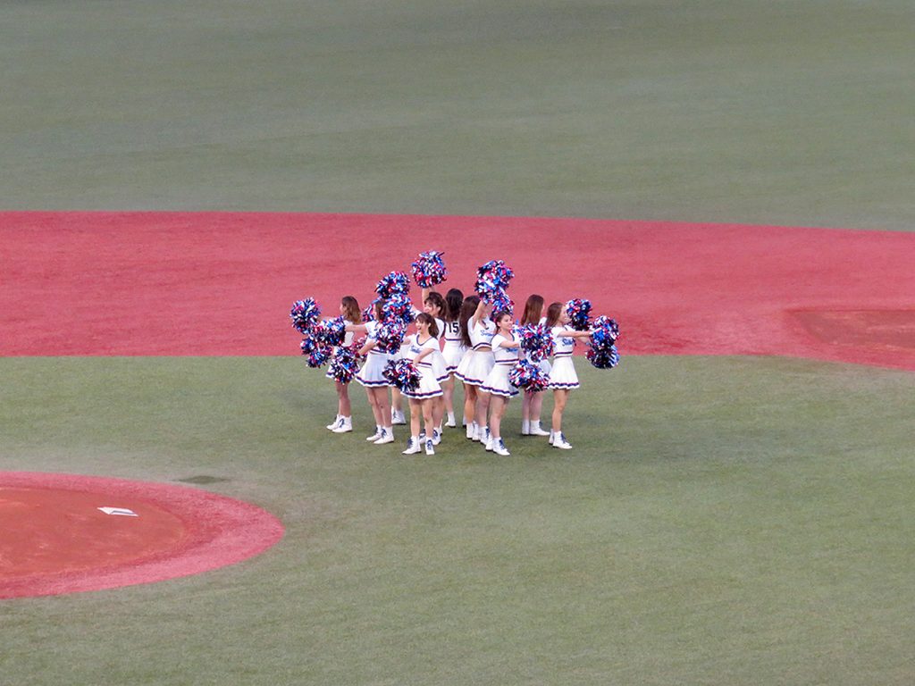 A group of young women wearing cheerleader outfits gather in a circle behind the pitcher's mound at a Tokyo Yakult Swallows baseball game.