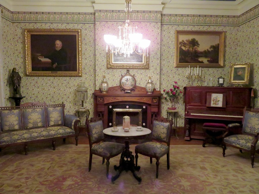 A glass chandelier in the center of a room above a small table with two chairs around it. A fireplace is along the wall in the background with a couch to the left and a piano to the right.