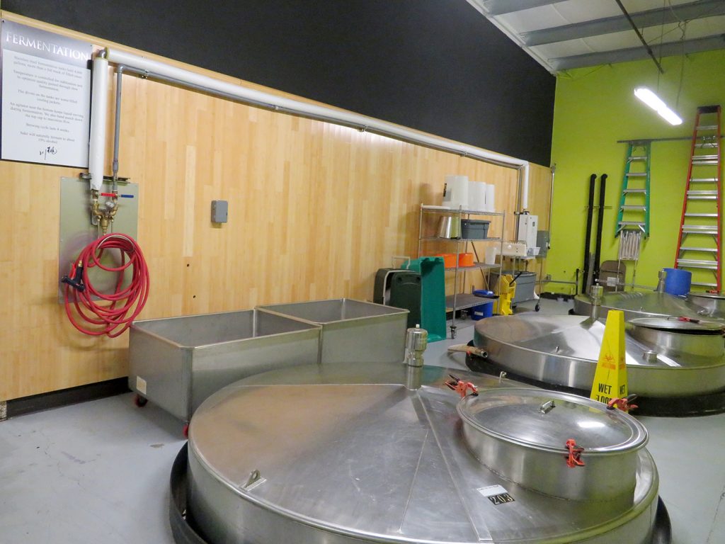 Overview of several large stainless steel tanks with a sign to the left on a wall that says "Fermentation," which includes information about the fermentation of sake.