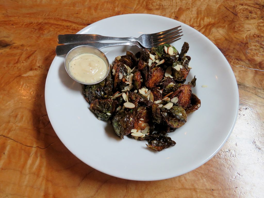 A circular white plate with grilled Brussels sprouts and a ramekin of garlic aioli on a natural wood table.