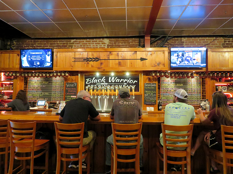 A large wooden bar with multiple chairs sitting at it with a wall of twelve tap handles in the middle and "Black Warrior Brewing Company" behind them.