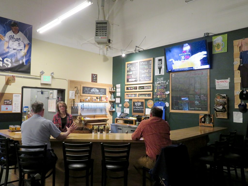 Overview of a large bar in a corner with people sitting at the bar and a server behind it. A sign above the tap wall says "Vertigo Brewing."