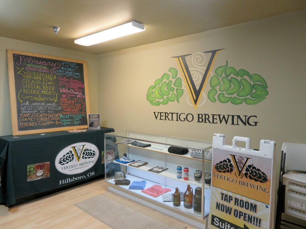 Table to left along with a large chalkboard on top of it. "Vertigo Brewing" is written on a wall to the right.